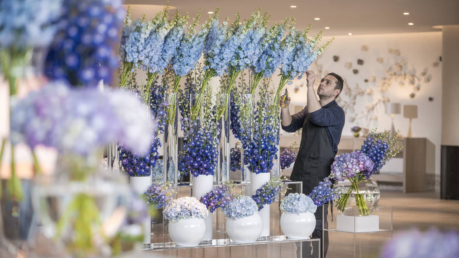 Florist tends to tall blue floral arrangements in glass vases in studio