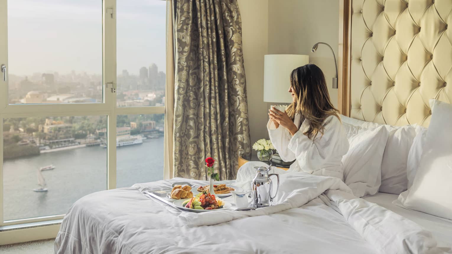 Woman sitting on a bed with a tray of food, looking toward the Nile River and Cairo from the window