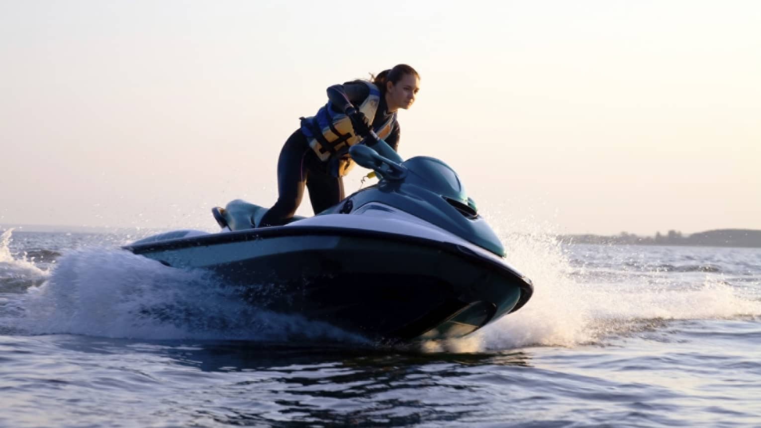 Woman wearing life jacket stands, steers jet ski on ocean at sunset