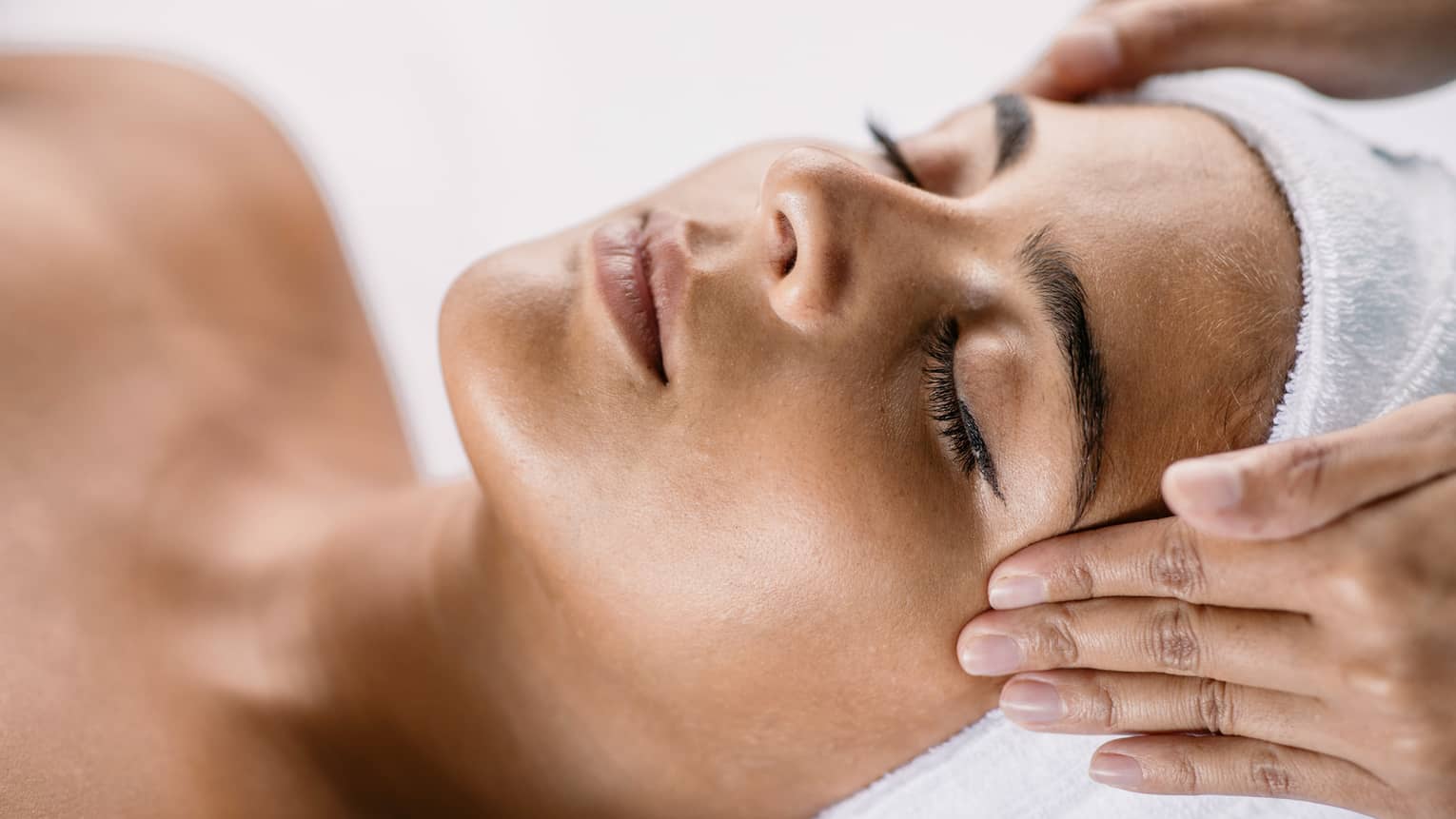 Hands massage woman's temples as she closes her eyes, spa towel around hair