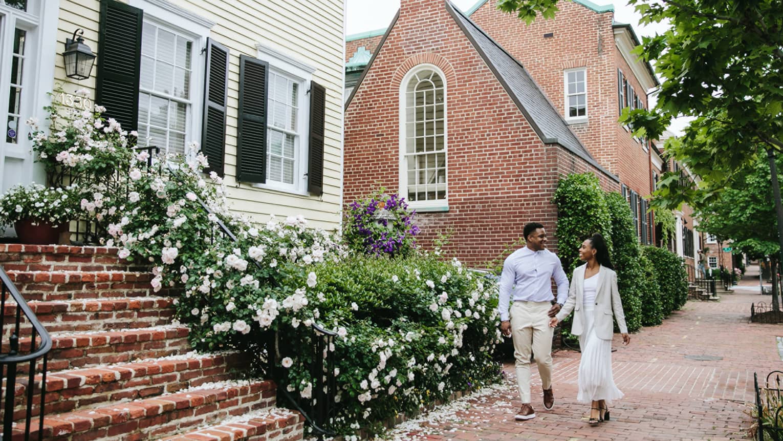 Two guests walking on a brick sidewalk beside houses and flowers.