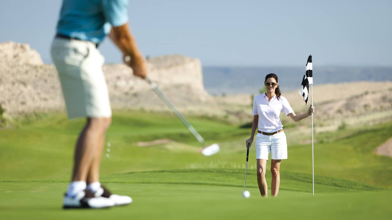 Woman holds golf flag near hole as man putts on course green