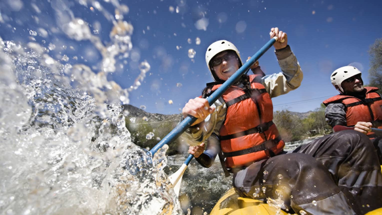 Smiling woman wearing red life vest holds paddles as waves crash over raft