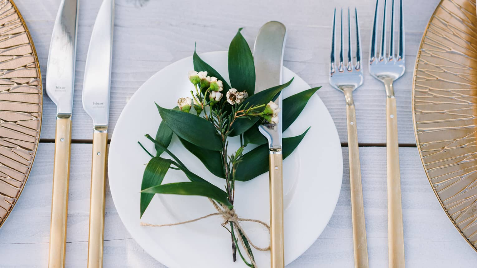 A bohemian table setting of gold chargers, modern long gold and silver flatware, white porcelain plates and a sprig of greenery
