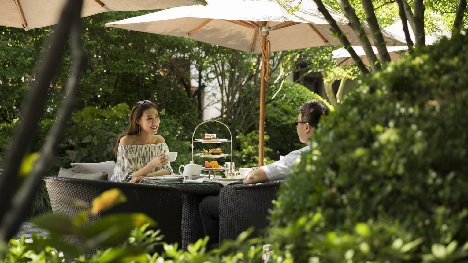 Woman and man drink tea near tray of sweets at table under shade of patio umbrella