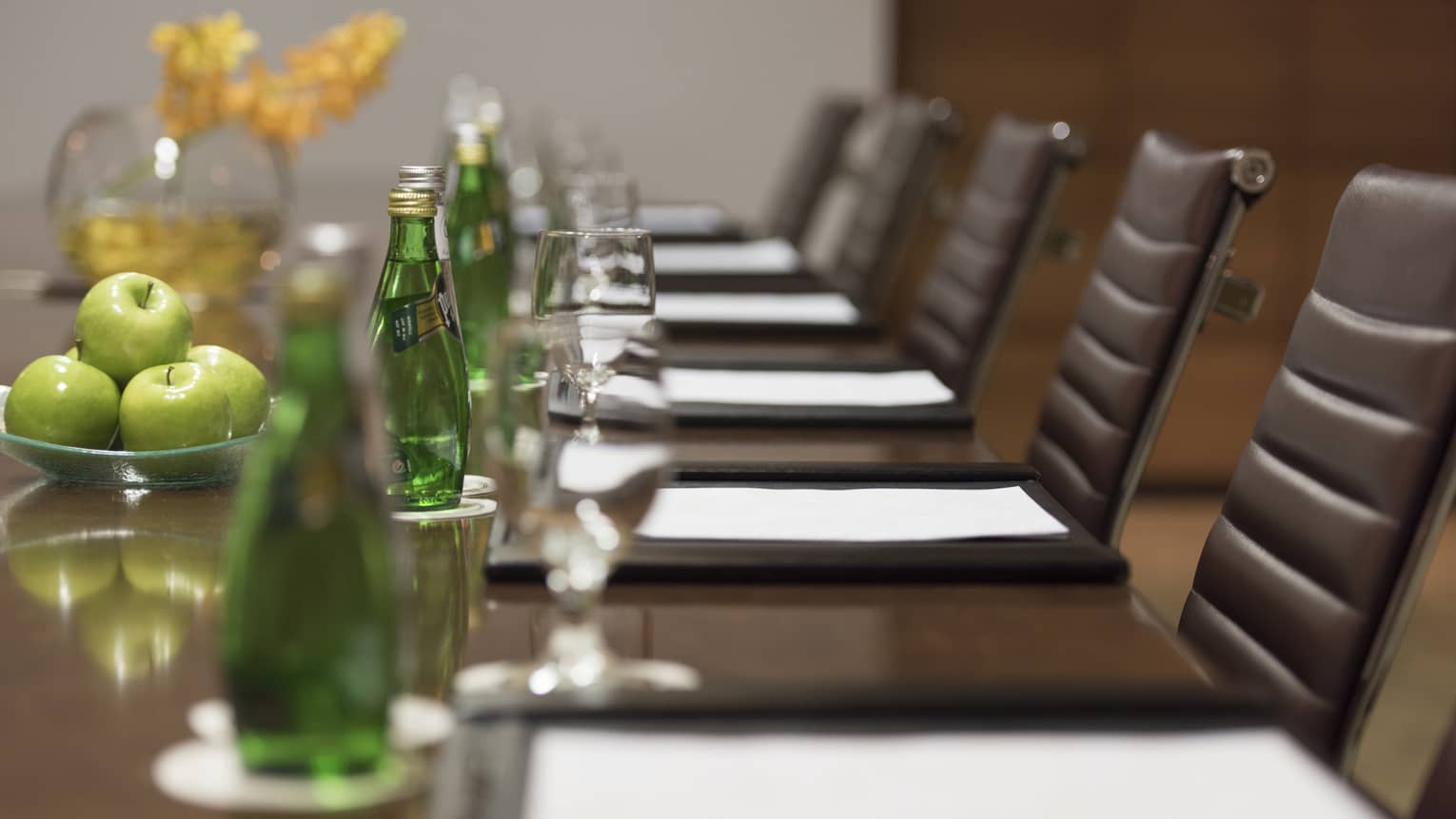 Close-up of business meeting table row of chairs, paper agendas, green glass bottles, apples