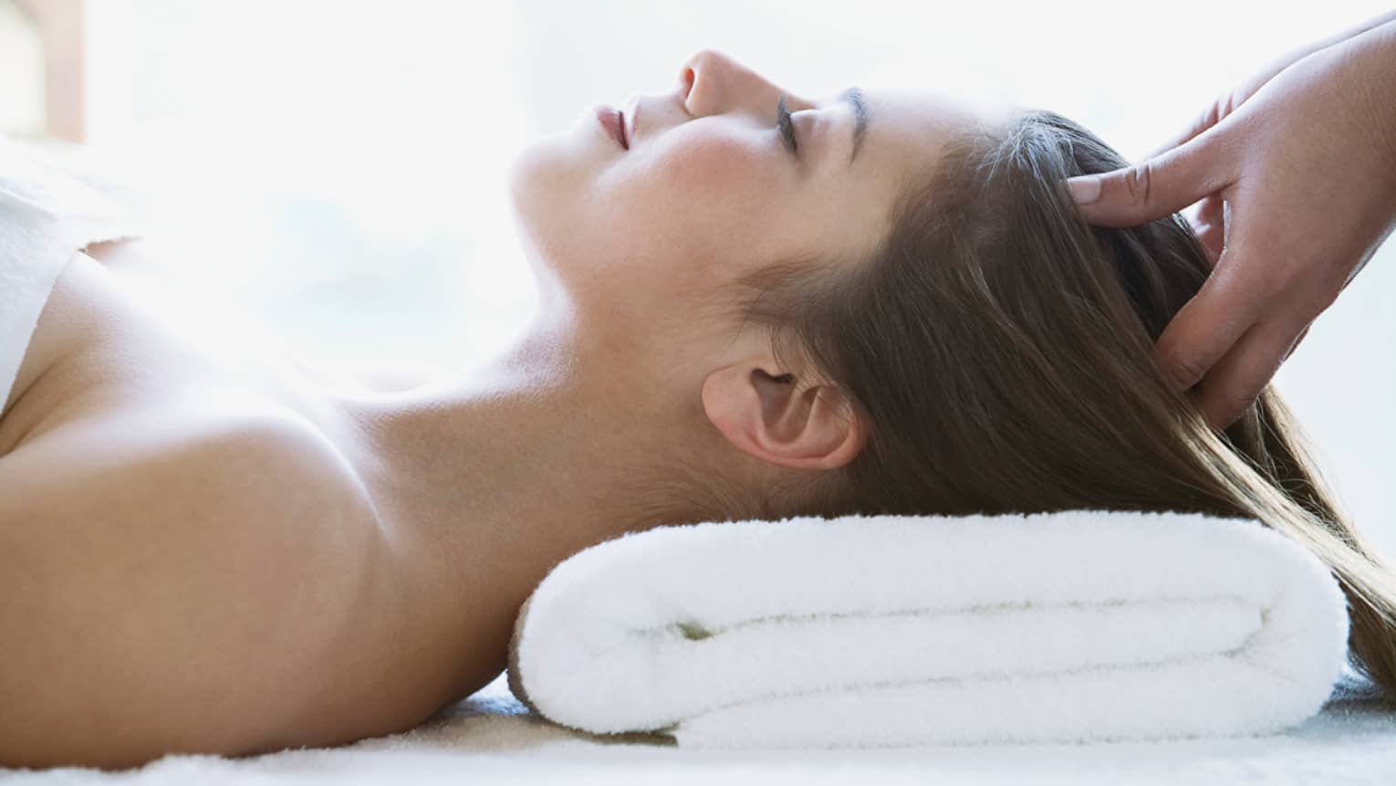 Woman lays on back with head on folded white towel, closes eyes as hands massage her scalp