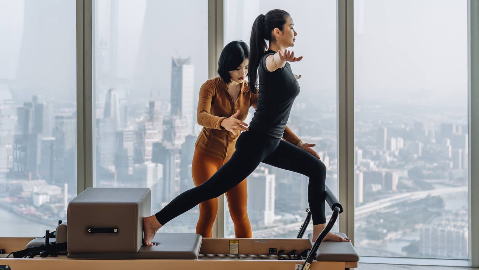 An instructor gently adjusts the positon of a guest in a lunge on a reformer machine, high up overlooking a hazy cityscape.