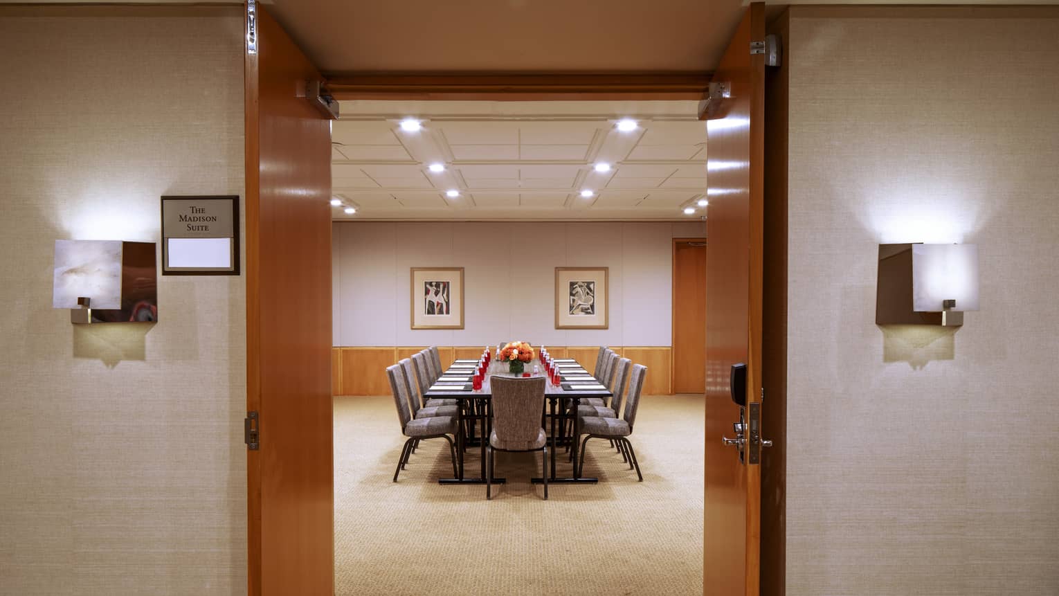 Two dark wooden doors are opened to reveal a function room with a long rectangular boardroom table with an orange floral arrangement in the center 