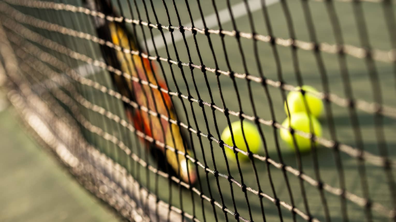 Close-up of tennis racket against net, three green tennis balls on ground