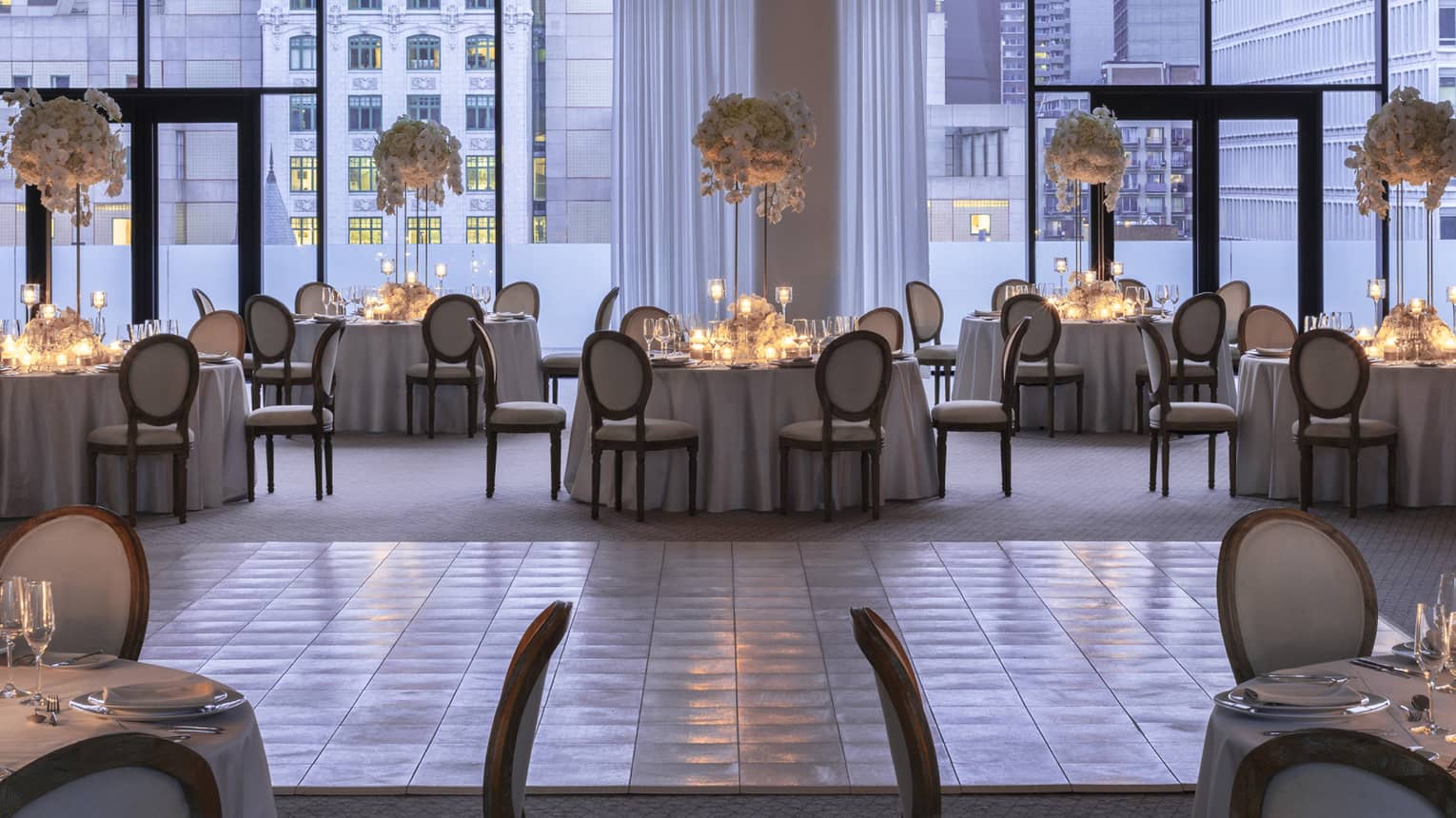 Ballroom with floor-to-ceiling windows overlooking buildings, round tables with candles and large floral displays 