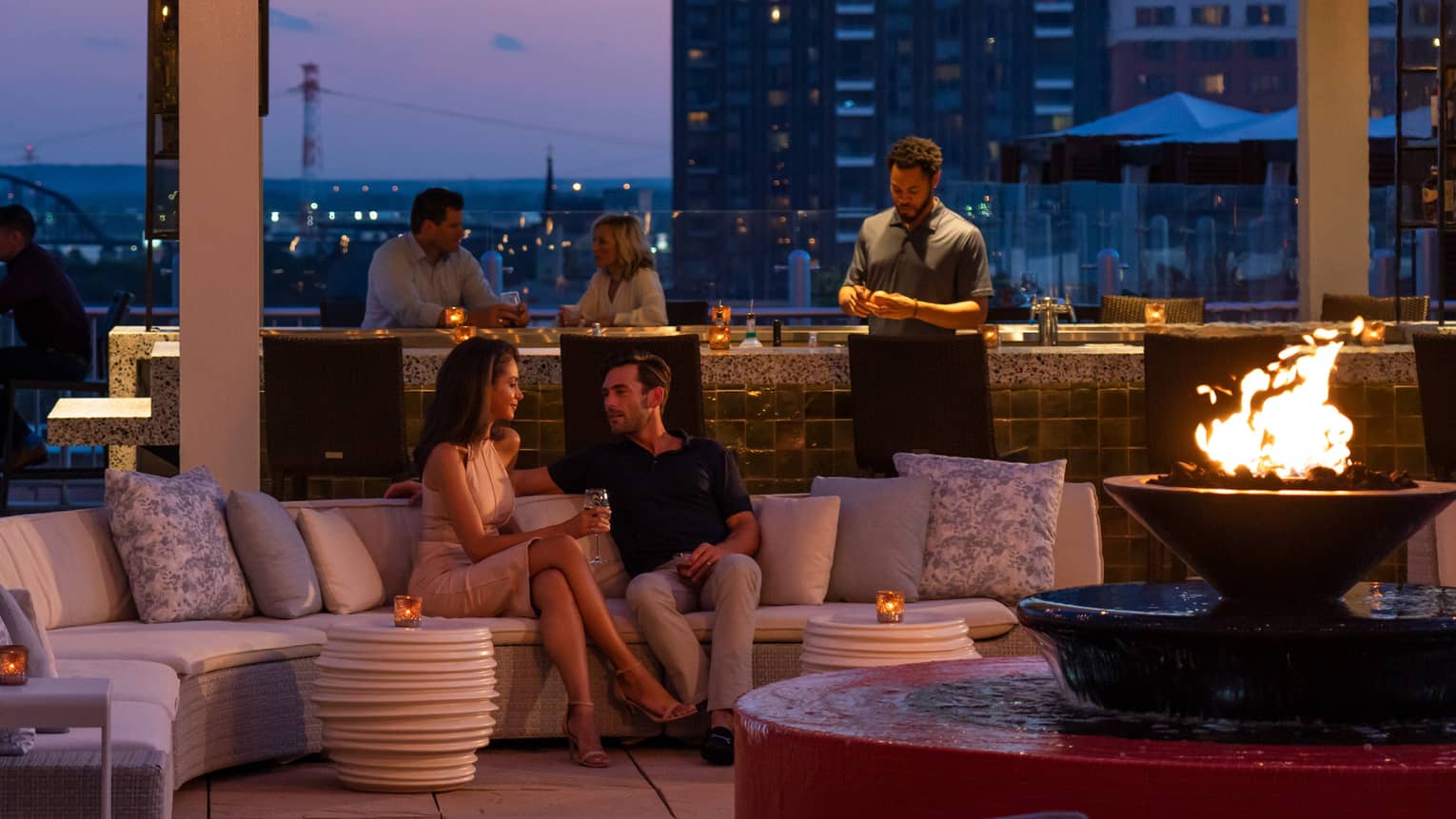 The sky is dark beyond the cinderhouse rooftop bar – furnished with a curved sectional, fire pit, hanging lights and people socialize 