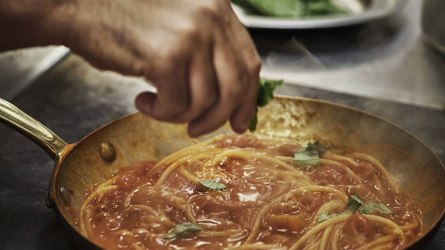 Fresh chopped basil is added to a steaming spaghetti dish in red sauce, prepared in a skillet on a stainless steel cooktop.