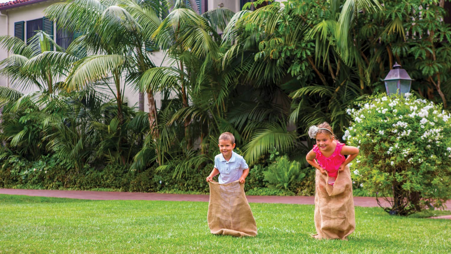 Two smiling children race across lawn in burlap sacks