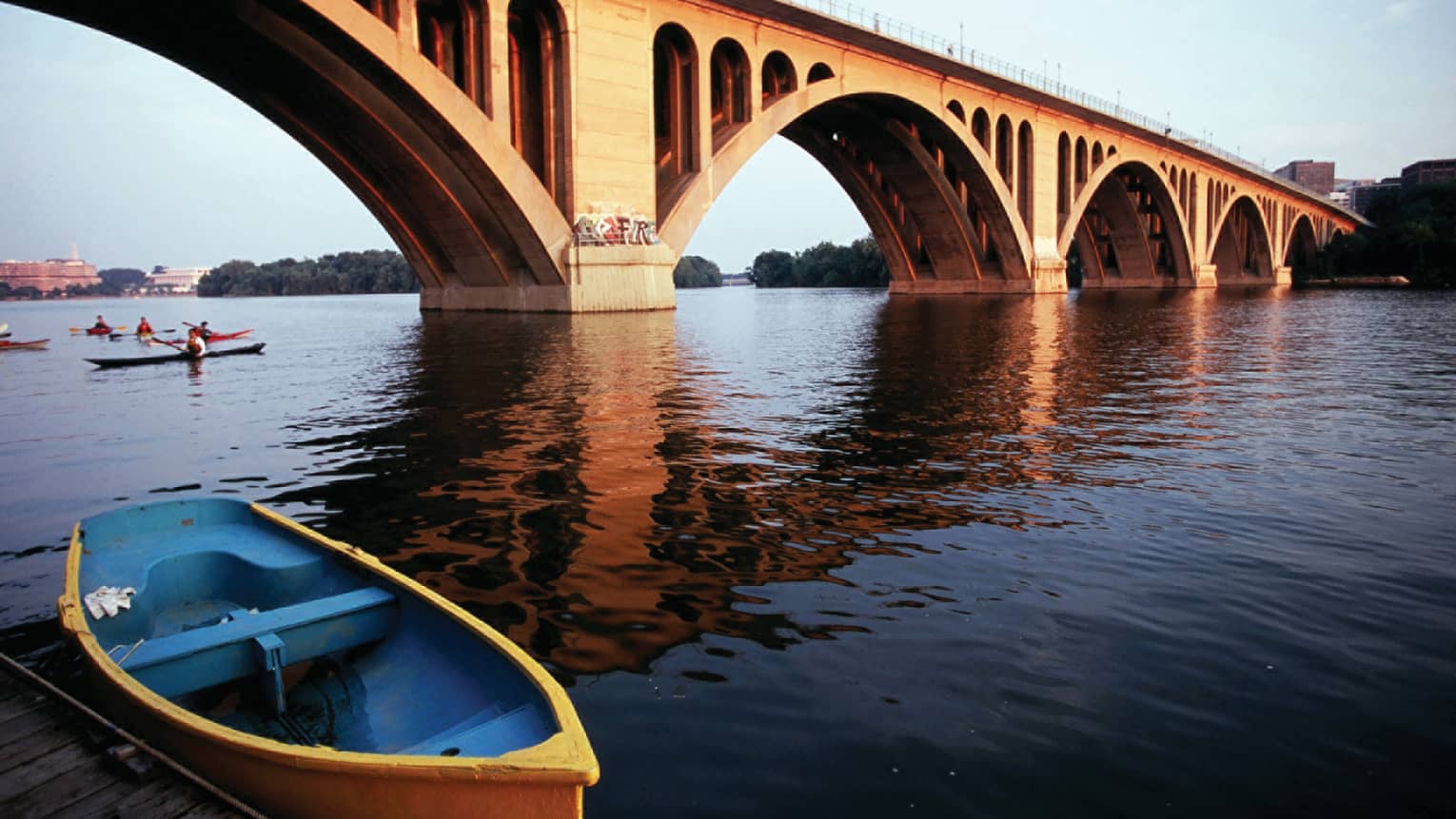 A wide river with a large bridge crossing it, there are people rowing kayaks under the bridge.