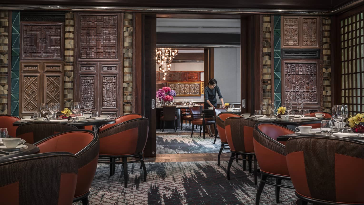 Woman sets table at Jiang-Nan Chun, Michelin-starred Cantonese restaurant, with red leather bucket chairs around dining tables, carved wood walls.