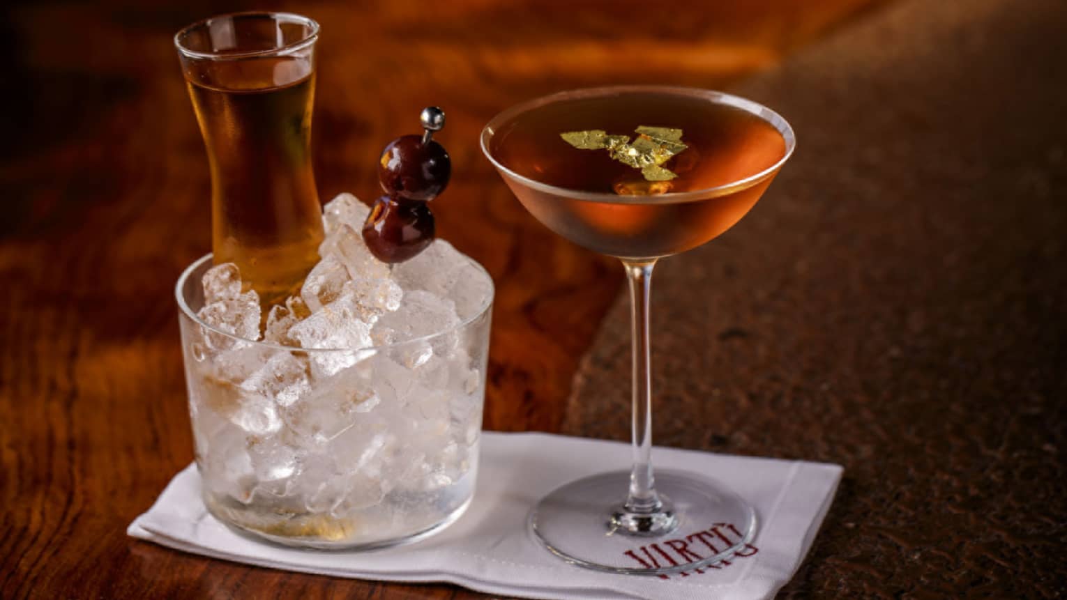 Two amber-coloured cocktails in stemmed glasses, one on ice with skewer of olives