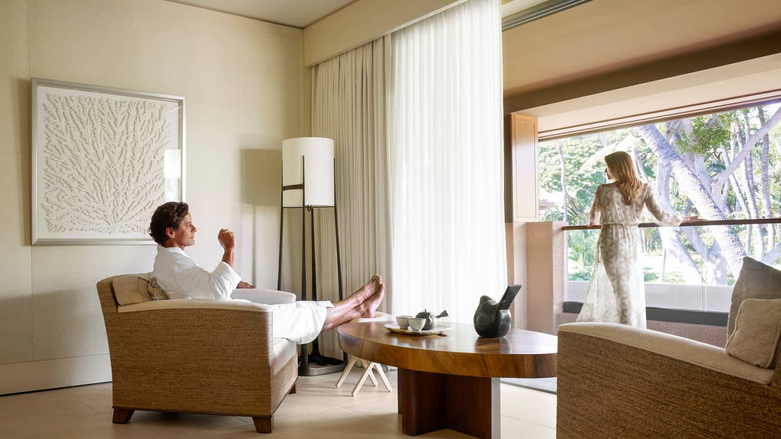 Man in white bathrobe kicks back in armchair in Spa room, woman in sheer robe stands at sunny window