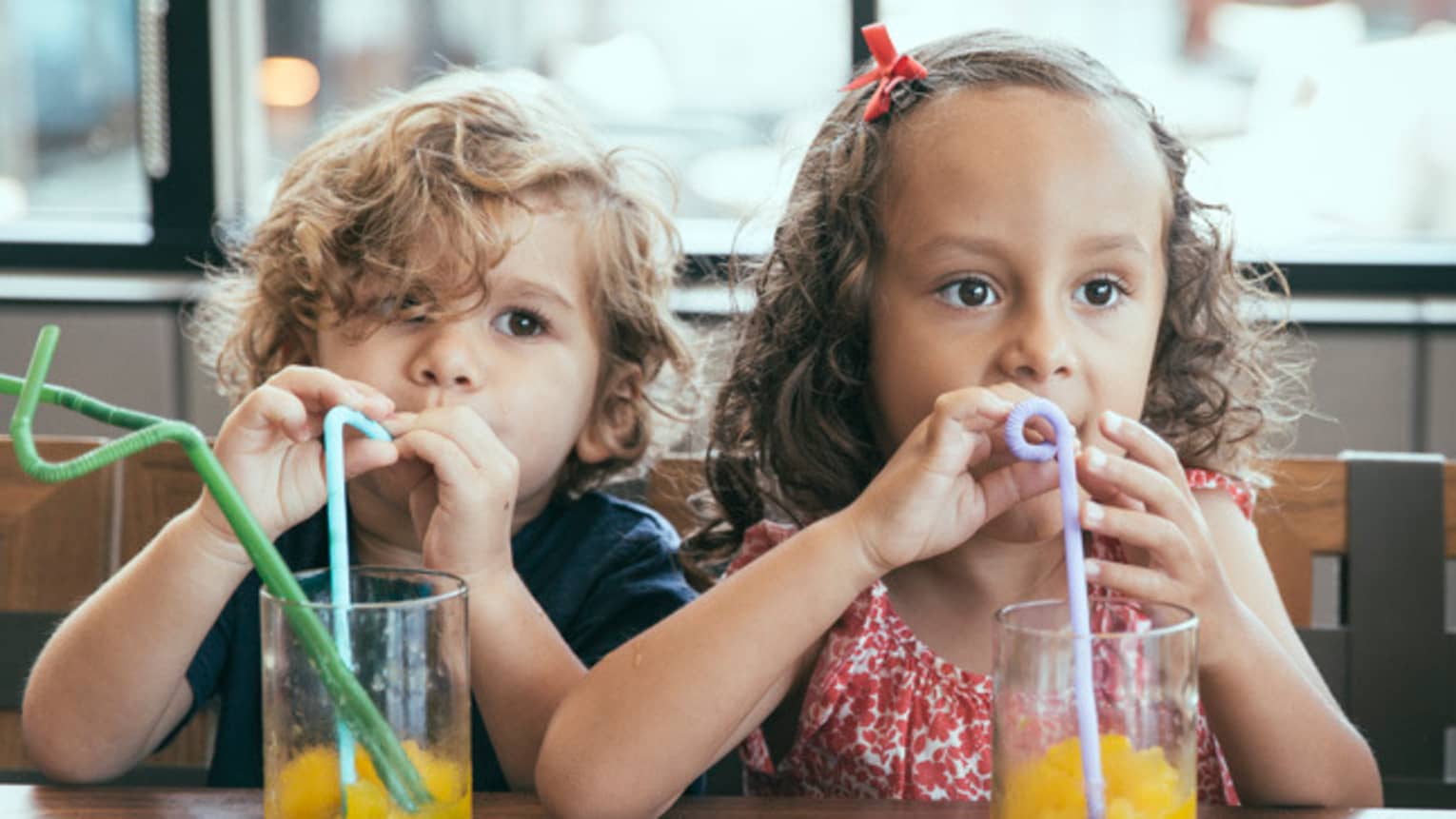 Two small children with curly hair sit side-by-side drinking juice with swirly straws at Club 760