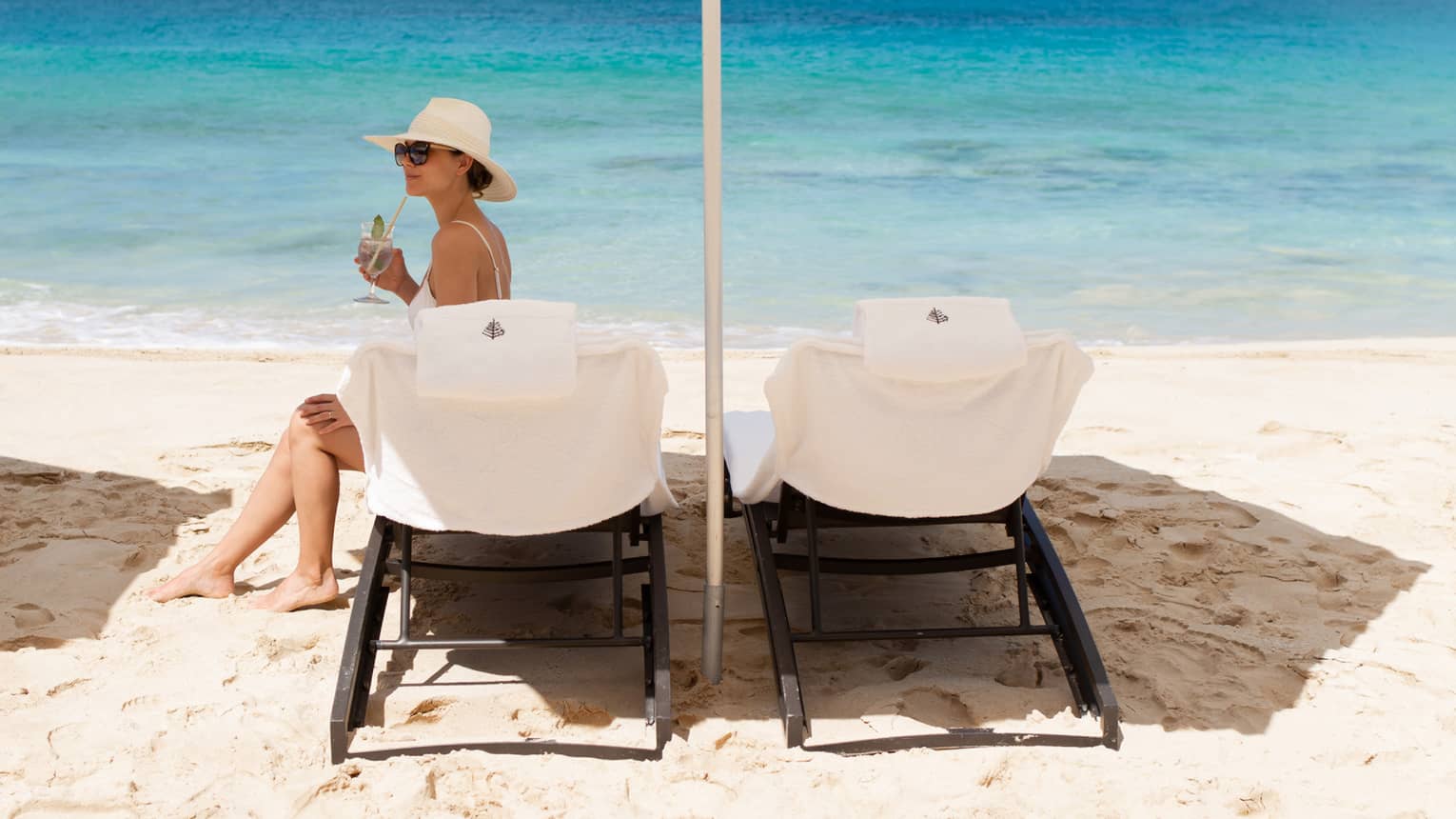 Two white beach lounge chairs a set up beneath a white umbrella on the sand near turquoise waters as a woman wearing a white swimsuit and sunhat sits on one of the chairs