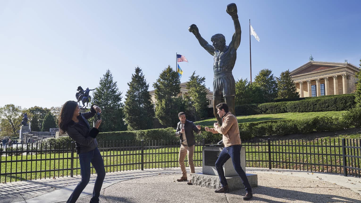 Two people pretend to box in front of the Rocky statue against a bright blue sky; another takes their photo, laughing.