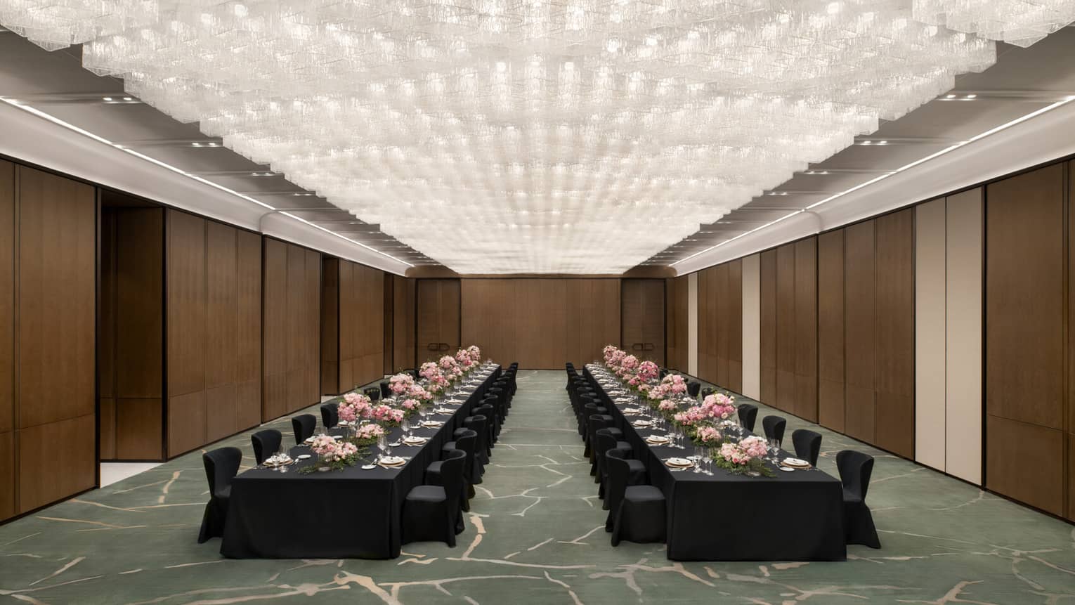 A spacious ballroom with two long black banquet tables set with plates, glassware, linens and pink floral arrangements