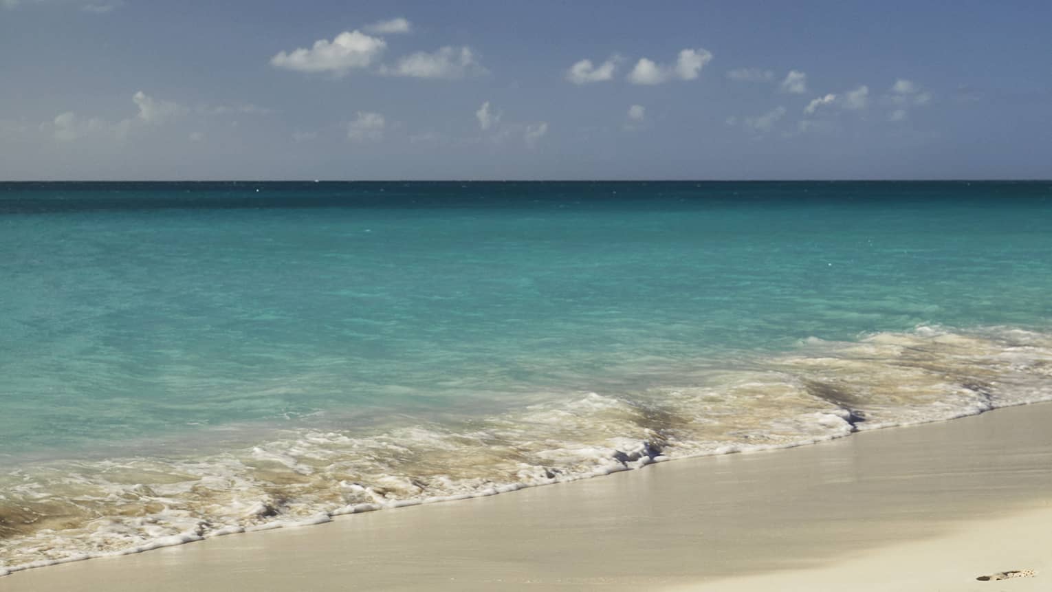 A sublime beachside view of endless turquoise ocean under blue sky dotted with wispy clouds, gentle waves lapping white sand.