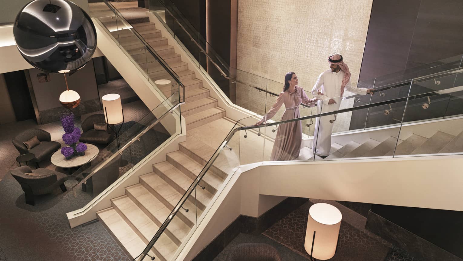 Couple ascends lobby stairs, cluster of armchairs and floor lamps visible below