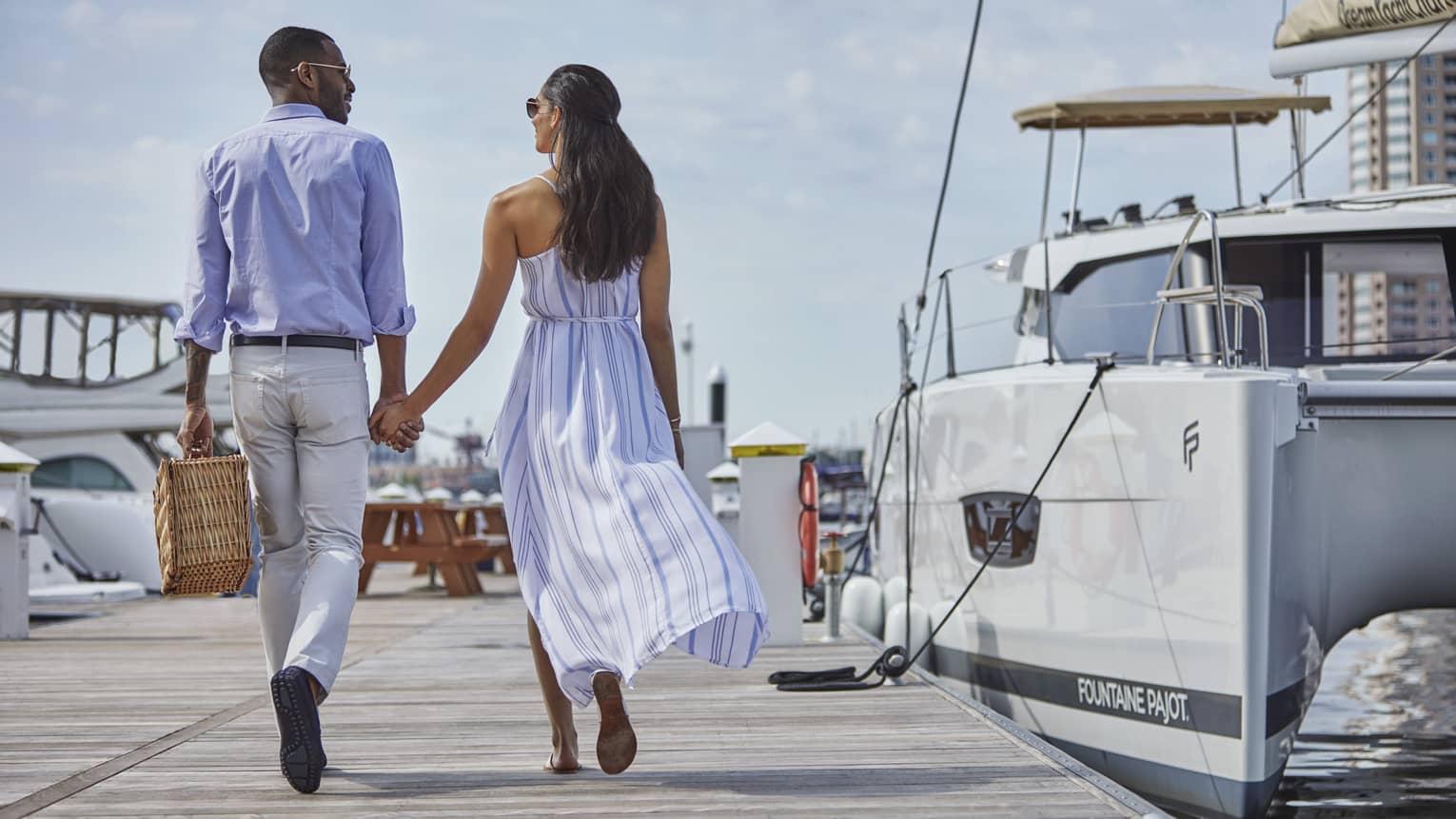Back of man and woman holding hands, walking along dock near sailboat