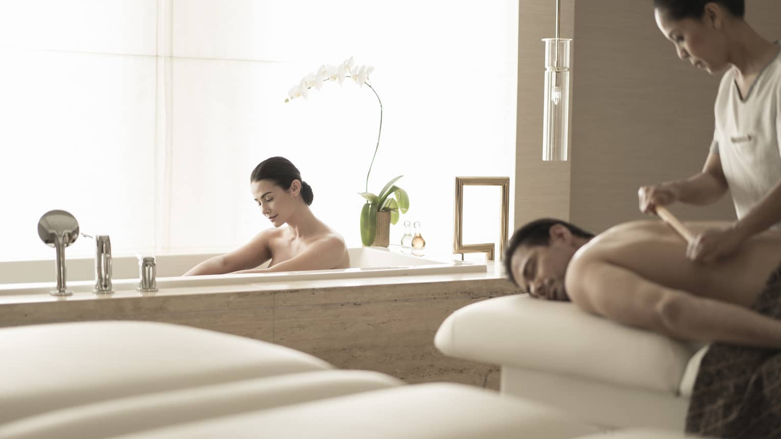 Woman soaks in spa tub near man getting massage 