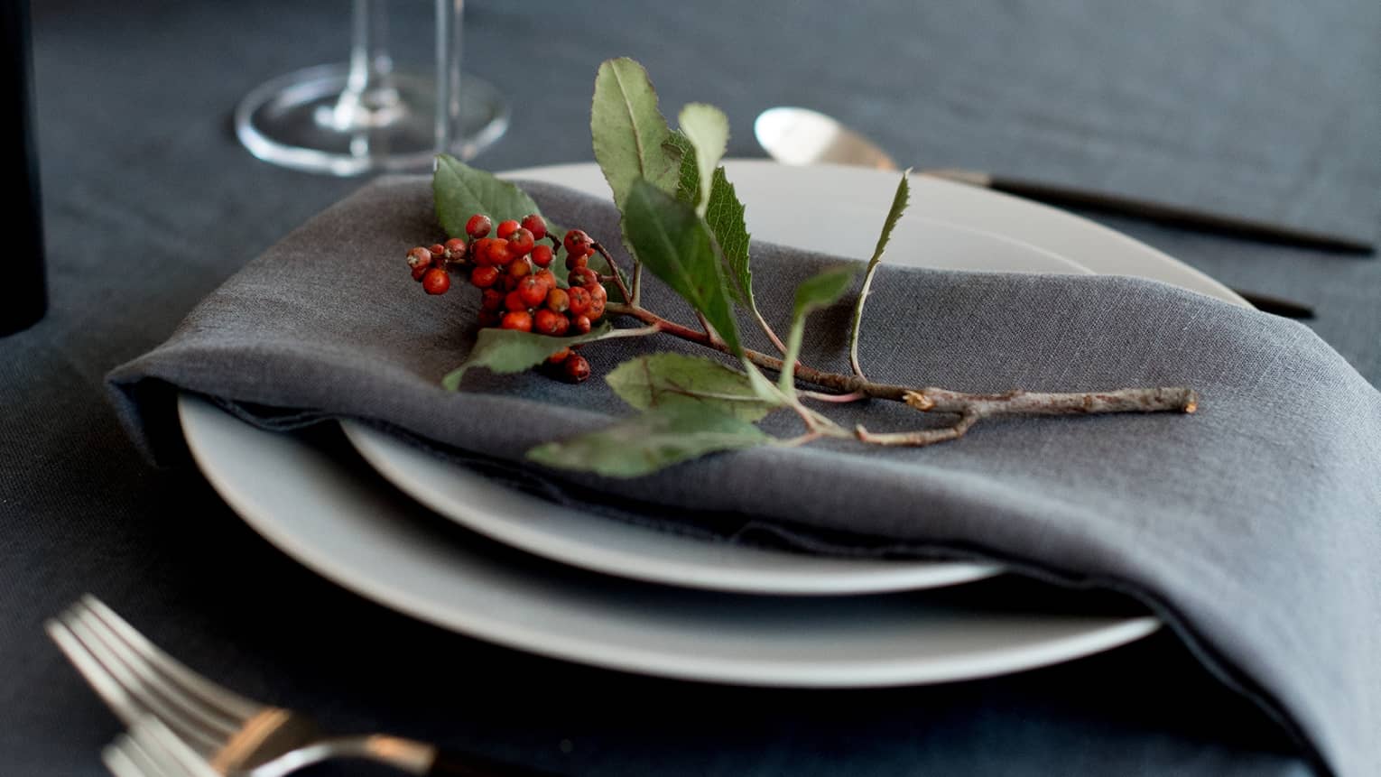 Table setting with white china topped with grey napkin and sprig of holly, cutlery, glasses