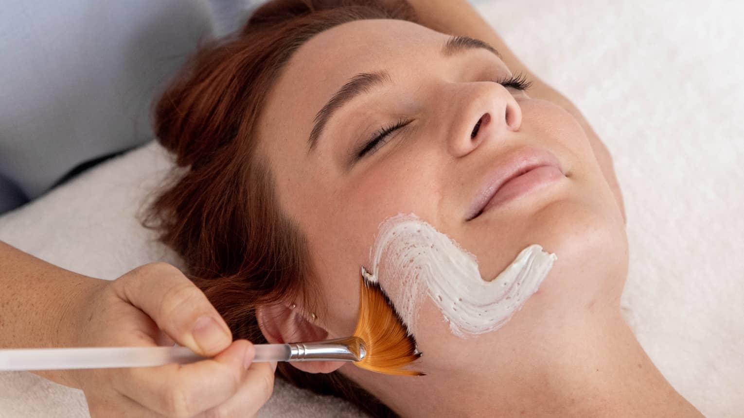 Spa staff paints white cream onto woman's face as she lays with eyes closed