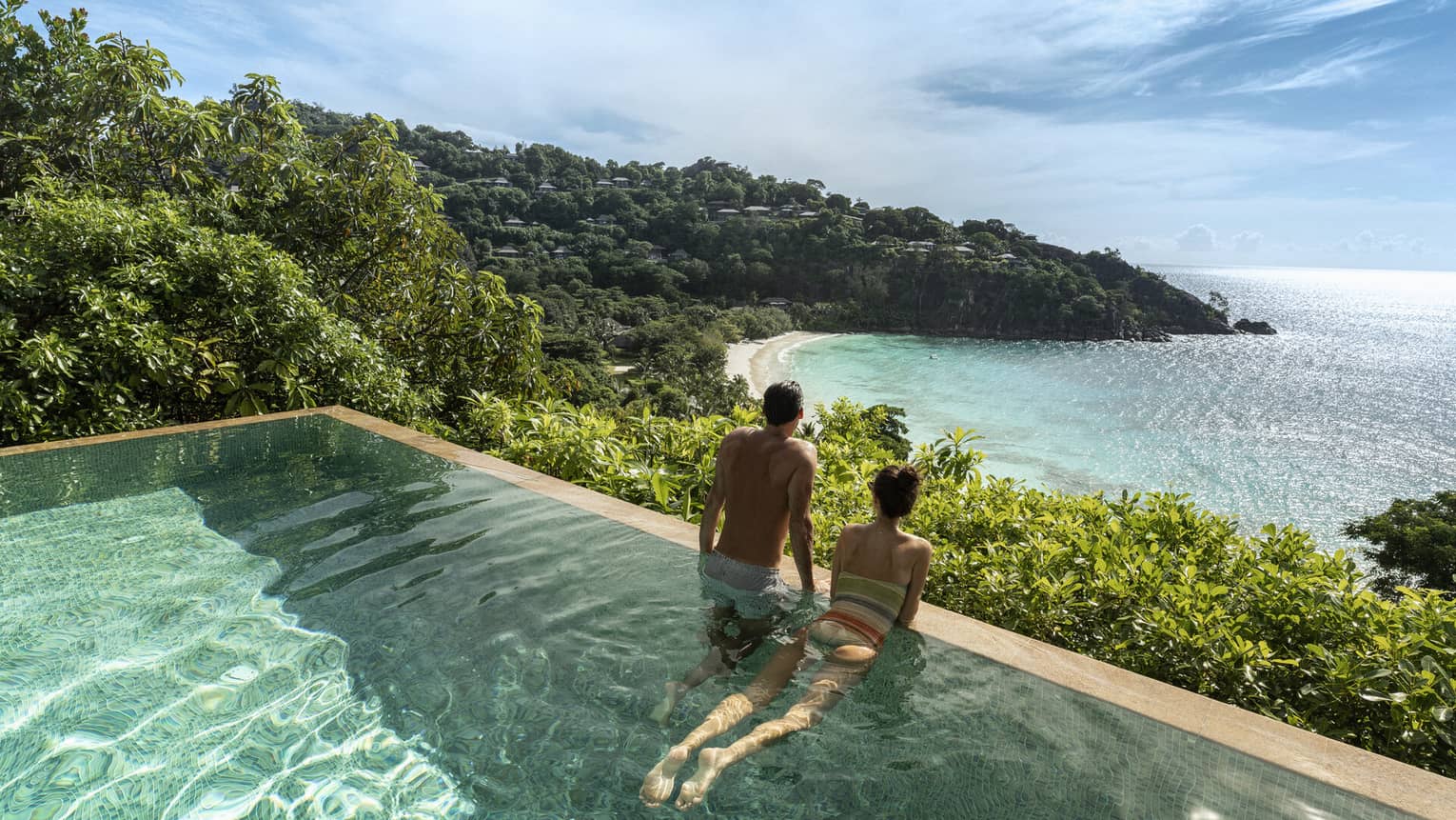 A couple looks out at a view of crystal blue waters and cliffs from an infinity pool