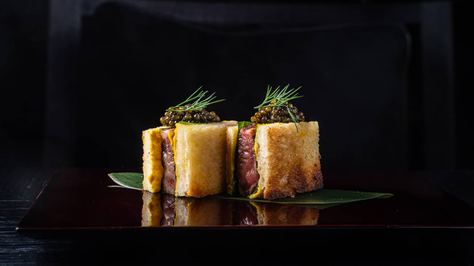 Sando Wagyu Beef topped with Caviar on banana leaf, dill frond garnish, black background