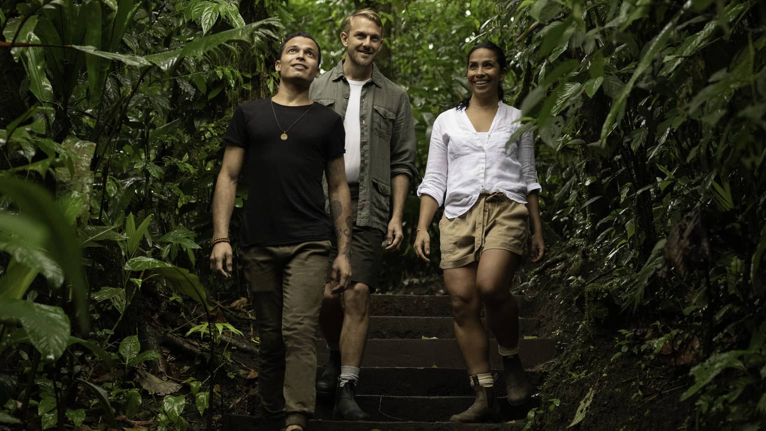 Three adventurous hikers smile as they walk down a wooden staircase surrounded by the lush green foliage of a rainforest.