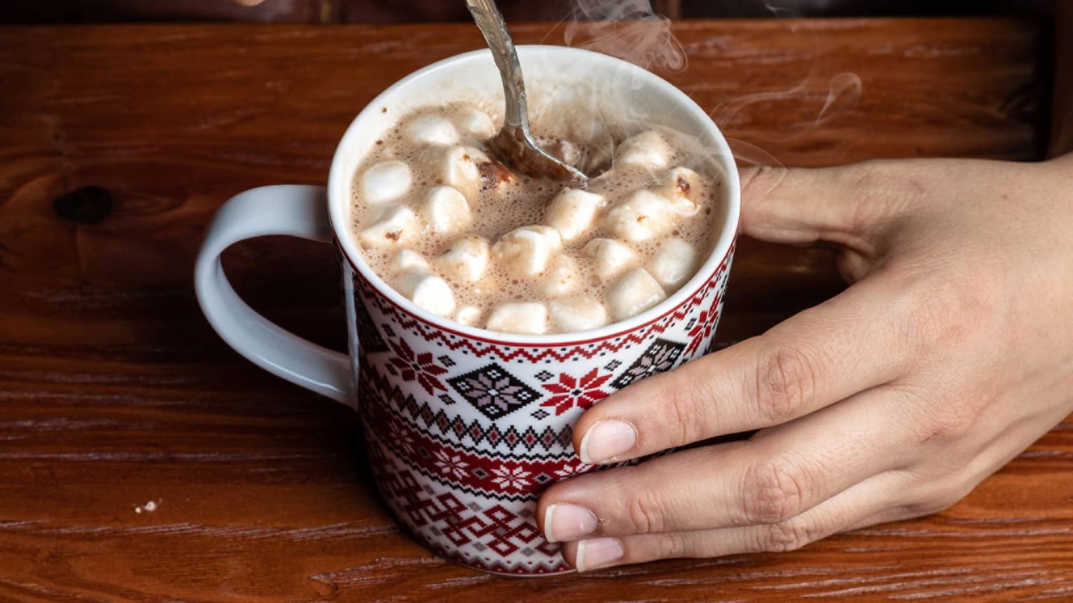 Close up of two hands holding and stirring hot chocolate with marshmallows in a patterned mug
