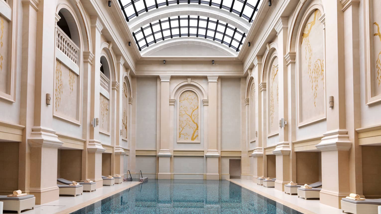 Long blue indoor swimming pool lined with deck chairs, tall cream-coloured panelled walls leading to curved sunroof 