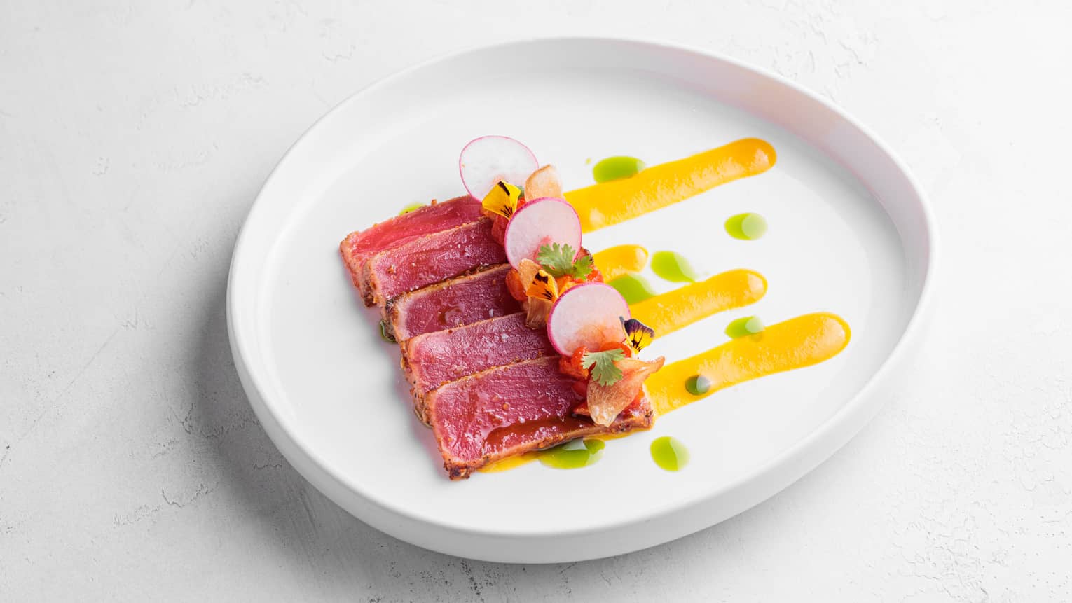 Five thin slices of rare tuna, sliced radishes, assorted garnish and thick lines of yellow sauce on a white plate