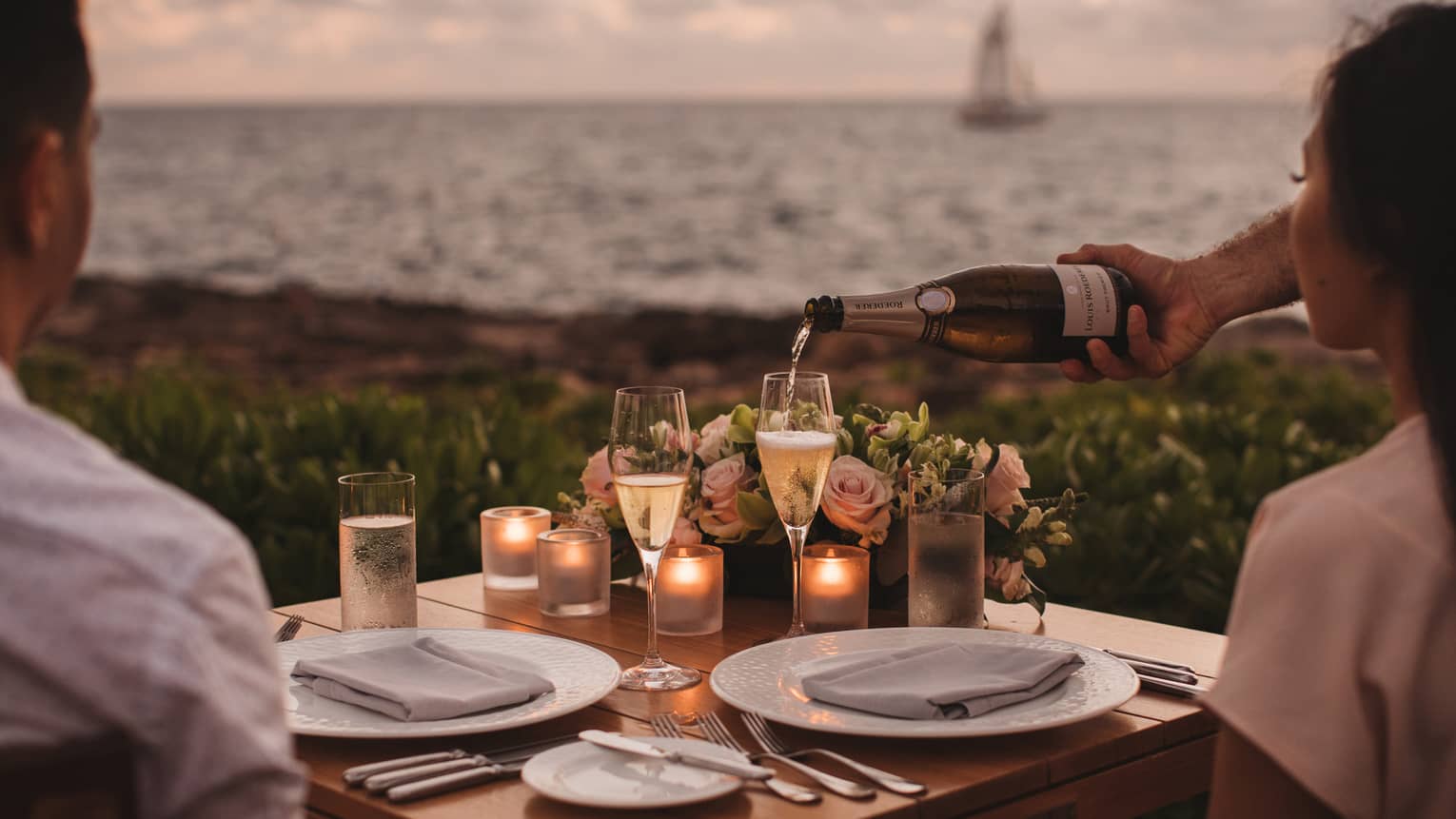 A couple having a private dinner on a coast with a sailboat in the distance.