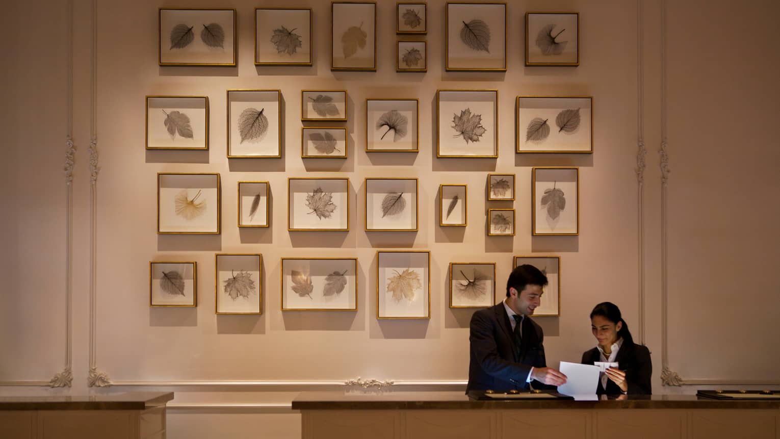 Two hotel staff look at paper at reception desk, tall wall with multiple framed leaf prints behind them