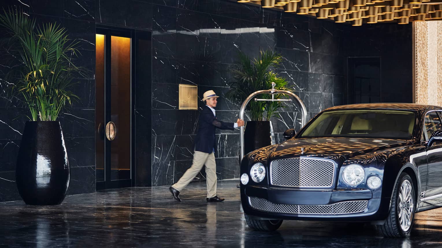 Man in suit jacket, fedora hat and khaki pants pushes cart near luxury car at hotel entrance 