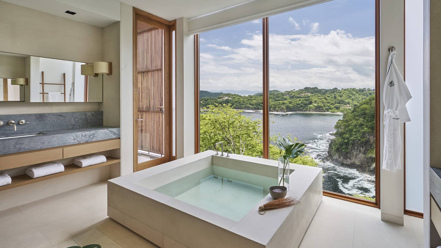 Bathroom with square stone tub; a wall of windows look out to the sea and trees