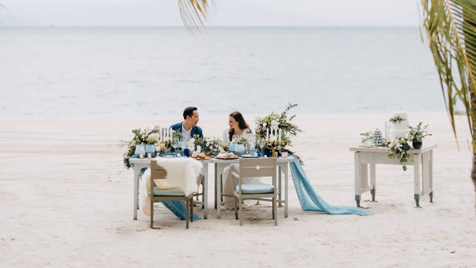 Smiling couple at private dining on white sand beach past palm leaves