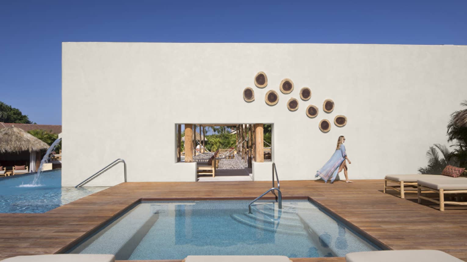 Woman wearing a long, flowy cover-up, walking past a stucco wall on a wooden pool deck