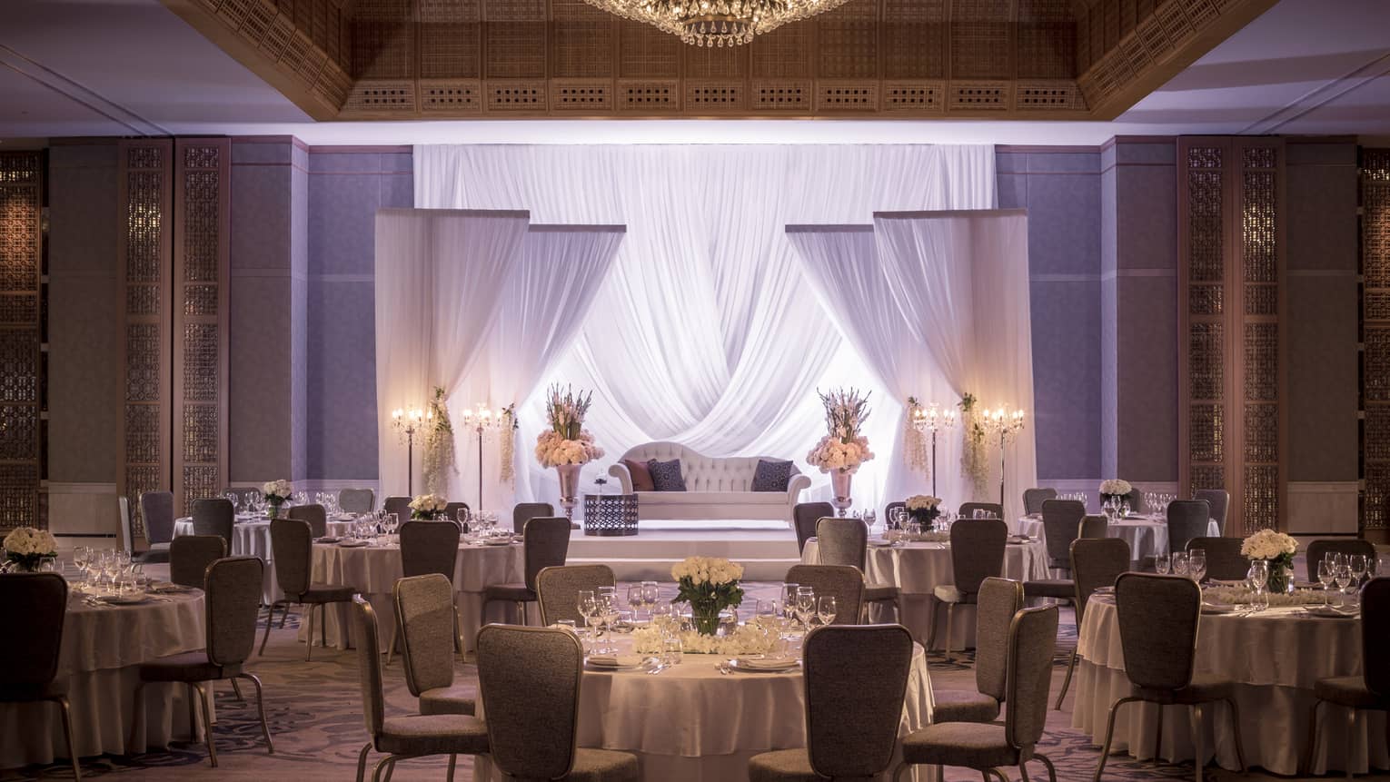 Round table and chairs facing podium with sheer curtains, flowers and chaise lounge in Jasmin Ballroom