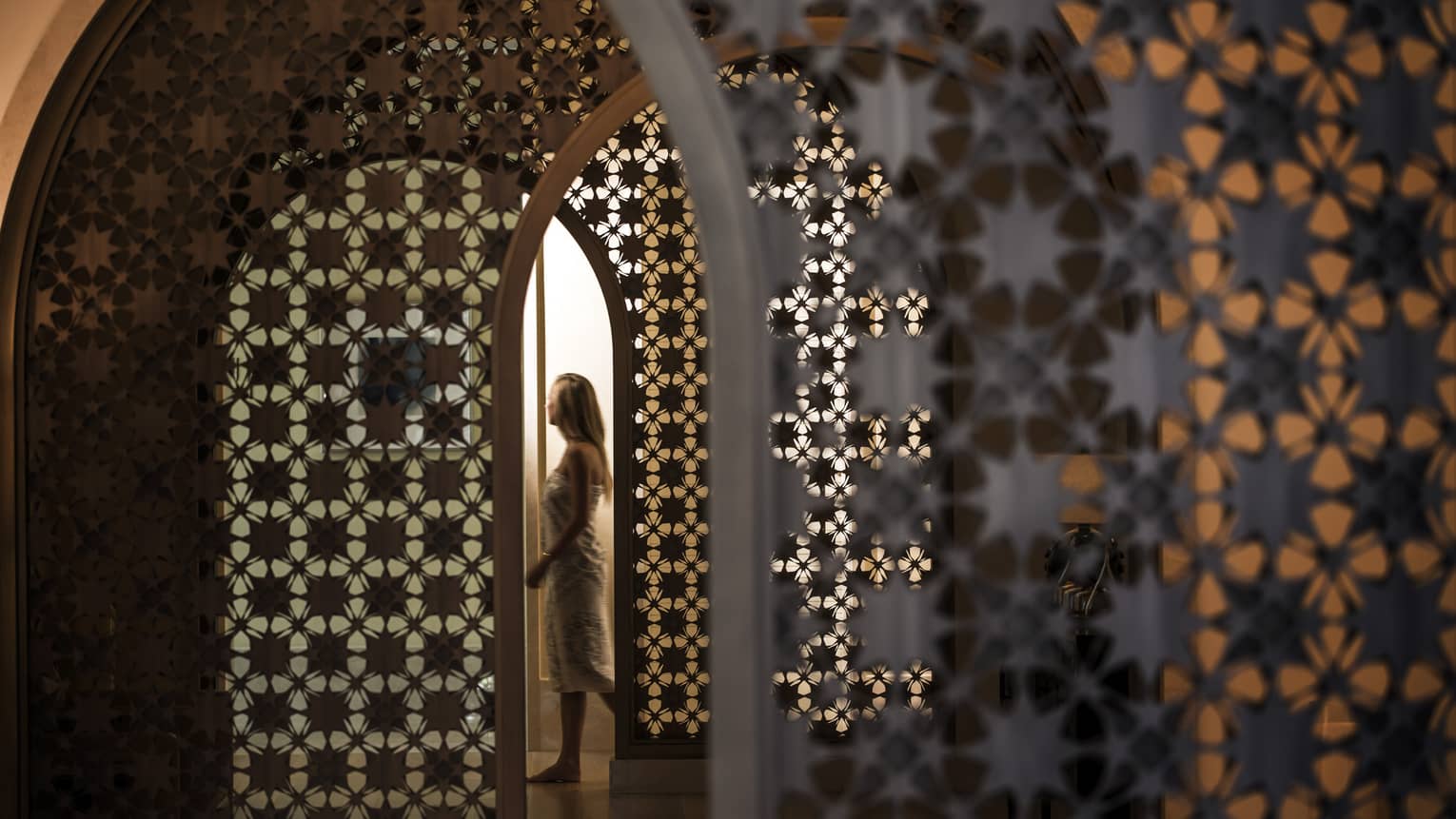 Woman in white wrap walks through dimly-lit decorative entranceway in hall