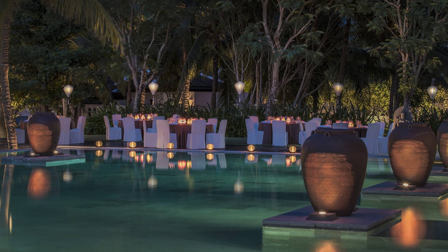 Tables and chairs with blue covers surround pool at night, surrounded by lanterns and vases