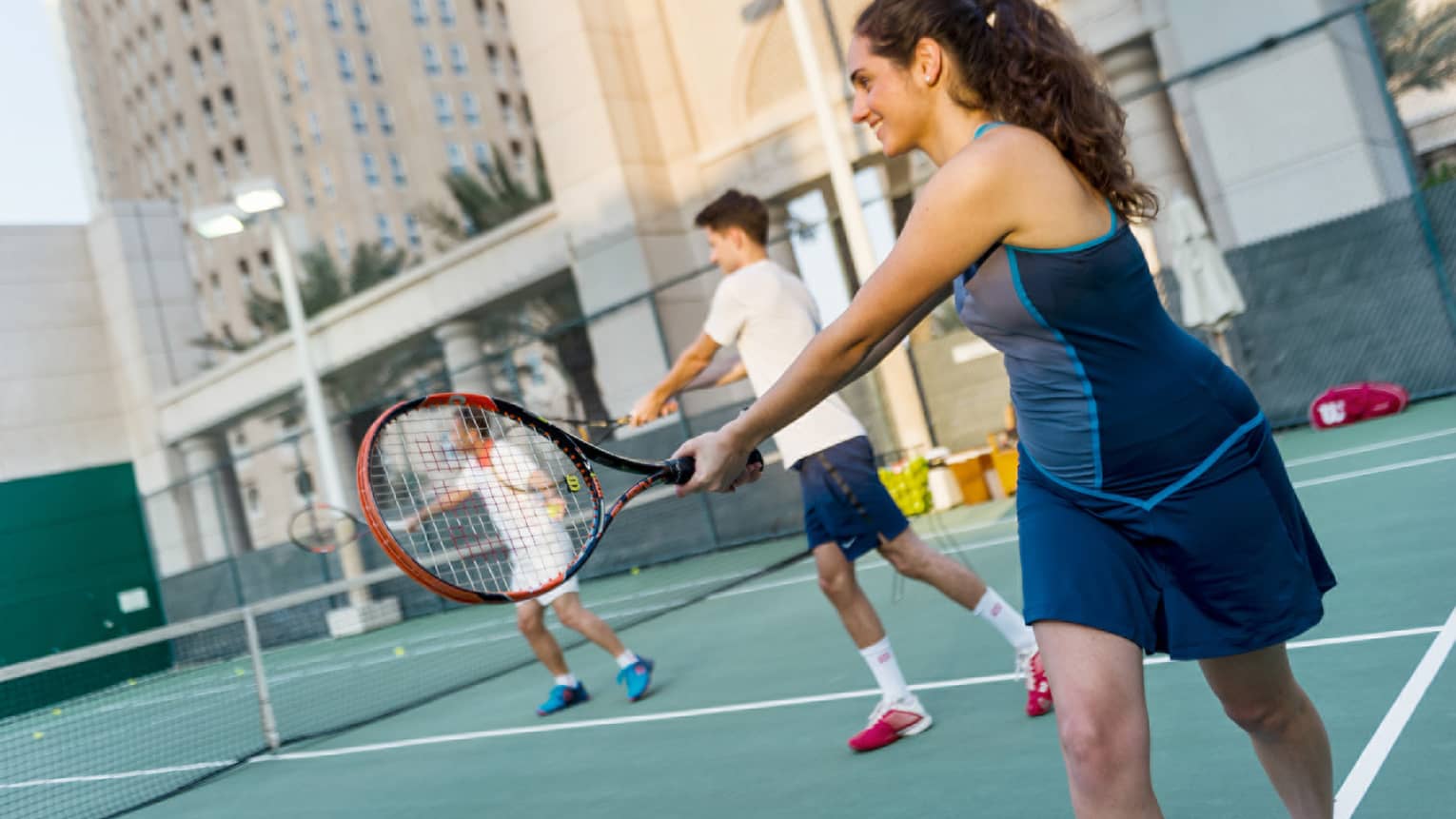 Woman in tennis outfit holds out racket, two people in background on court