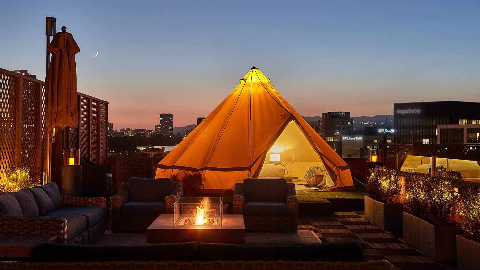 Close-up of illuminated white tent with bed, lamp by wicker chairs, modern outdoor fireplace