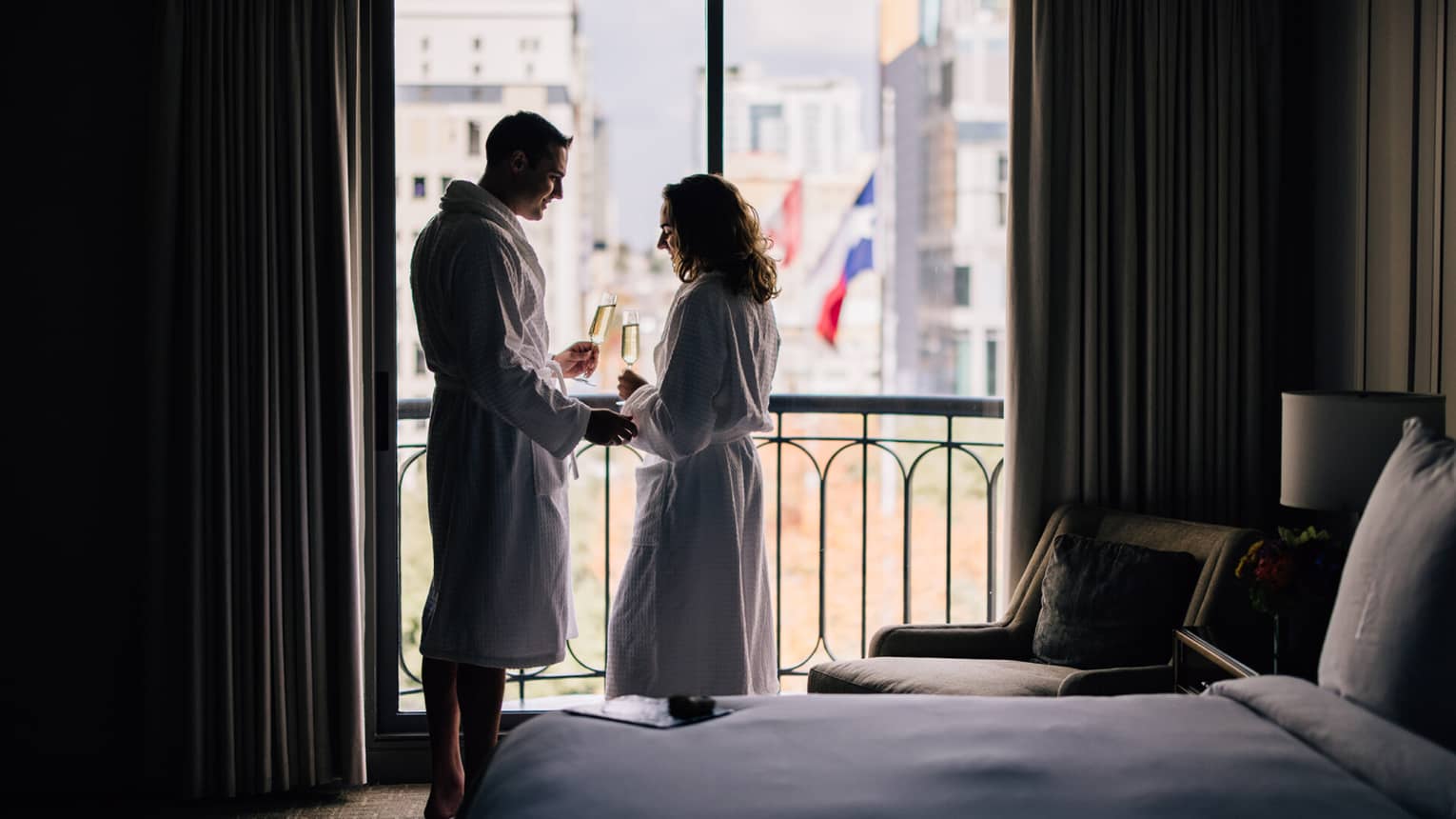A couple dressed in white bath robes toast champagne flutes in front of a balcony window, overlooking downtown Austin, Texas
